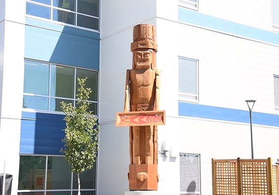 A wooden pole, shaped like a man, stands in front of a new, modern building that is blue and white