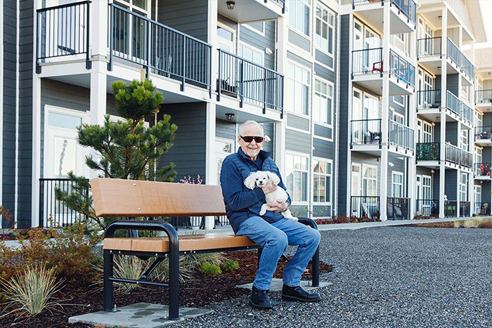 An older man with white hair sits on a bench holding a dog in front of a modern apartment building