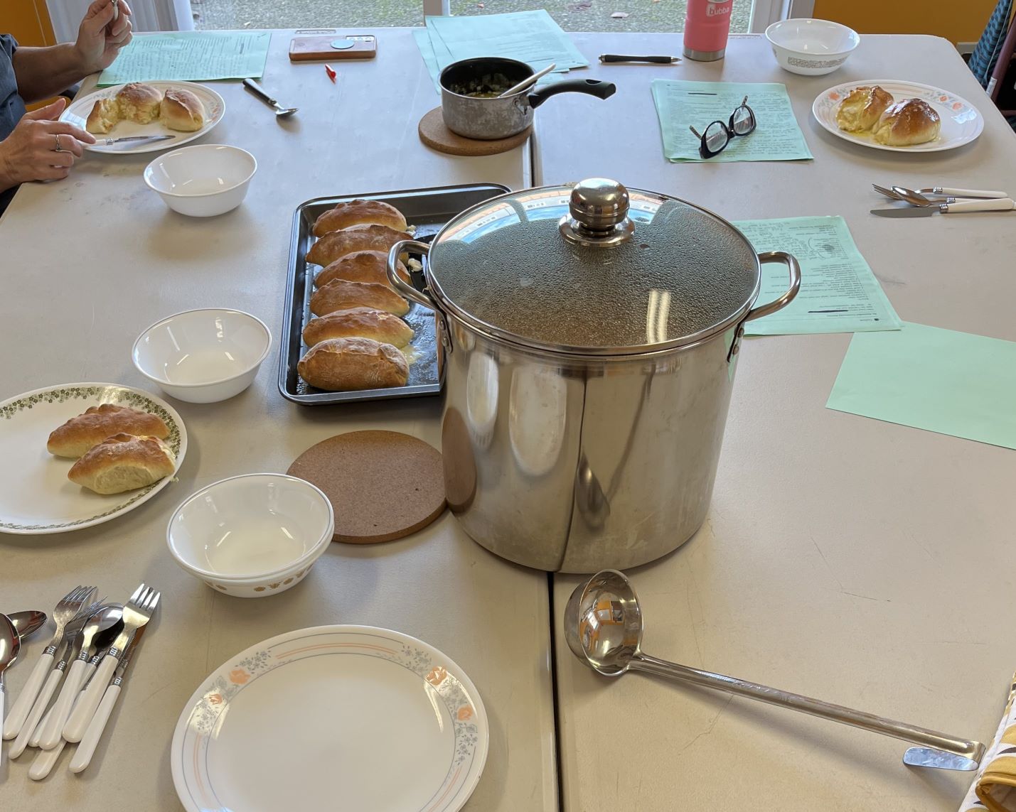 Table set for a shared meal with a large covered pot, ladle, plates, bowls, cutlery, and a tray of bread rolls