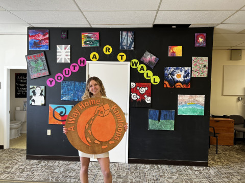 A young white woman stands in front of a youth artwork wall, holding her own artwork that reads “A Way Home, Kamloops” and features footsteps leading to a home.