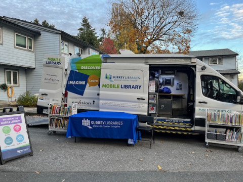 A white van with its sliding door open, full of books. A table with a blue tablecloth and two trolleys filled with books sits outside in front of the van.