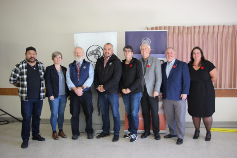 A group of people stand in a line, smiling in front of Huu-ay-aht branded promotional boards.