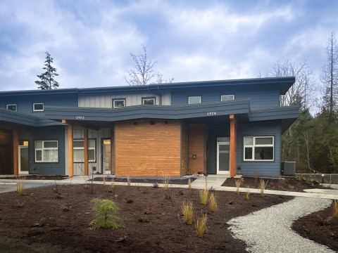 A modern, single-story duplex with blue and wooden panelling, large windows, and slanted roofs, surrounded by newly landscaped grounds with a winding gravel pathway leading to the entrances.