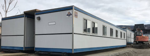 White with blue trim portables with 7 windows, lined up next to each other with a 4-foot hallway between them that has a wooden roof covering over it.