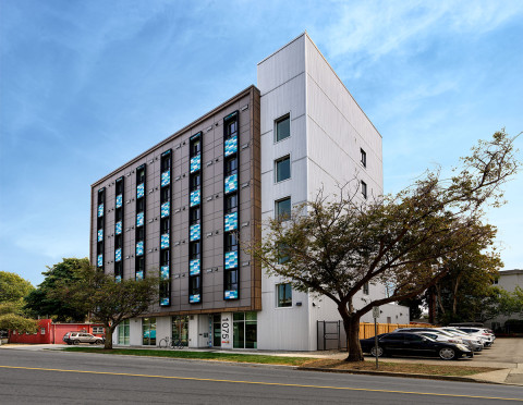 Six-storey modern apartment building at 1075 Meares Street, featuring a mix of brown and white exterior panels, vertical rows of blue-accented windows, and a glass entrance at street level. Trees line the sidewalk in front, and a small parking lot with cars is visible on the right.
