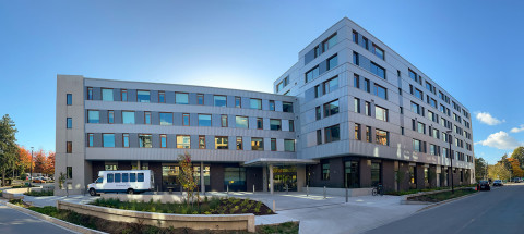 A modern six-storey building with grey and white exterior panels, large windows, and balconies, located at a landscaped street corner under a clear blue sky.