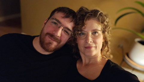 Young brown-haired man leaning next to his smiling, curly-haired mother.