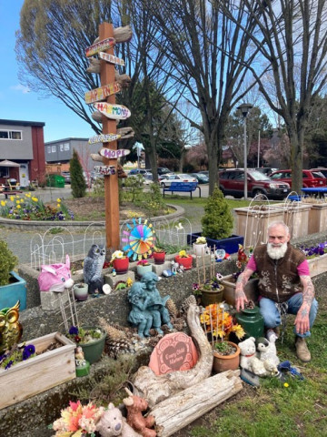 Older man with a long white beard sits in a community garden decorated with garden art, with a tall wooden post in the background.