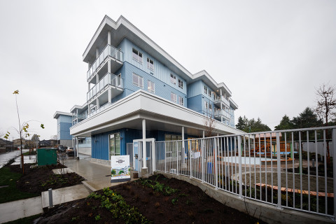 A modern, three-story building with light blue siding and white trim, featuring multiple balconies and large windows. The structure is surrounded by a metal fence, landscaped garden beds, and a paved walkway under an overcast sky.