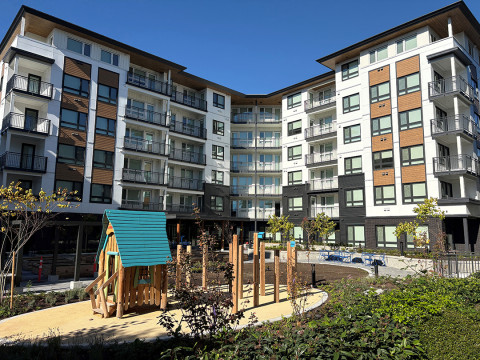 Modern apartment buildings with balconies overlooking a small playground and landscaped courtyard on a sunny day.