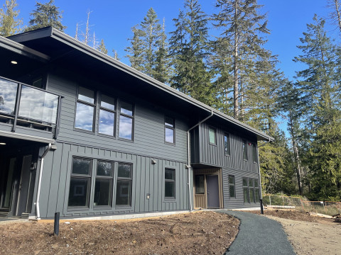 Modern two-story gray building with large windows, set among tall evergreen trees under a clear blue sky.