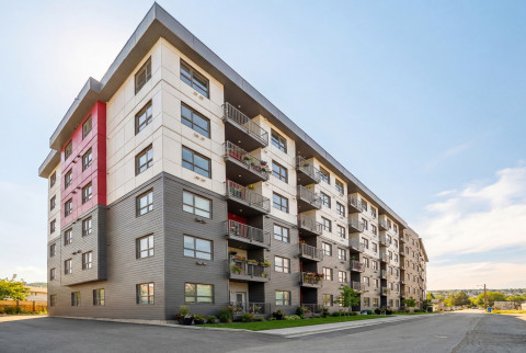 Modern multi-story apartment building with balconies and red accents beside a quiet street under a clear sky.