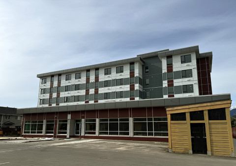 Modern four-story building with a flat roof, large ground-floor windows, and multicolored panel façade under a cloudy sky.