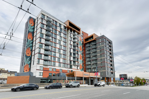 Modern mid‑rise building with balconies and colorful murals, set along a street with parked cars and overhead wires.