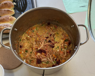 Large metal pot filled with a chunky soup sitting on a table beside a tray of bread rolls.