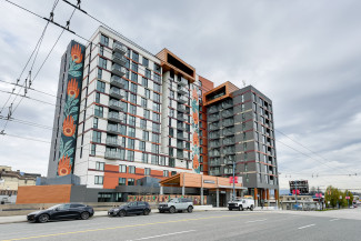 Modern mid‑rise building with balconies and colorful murals, set along a street with parked cars and overhead wires.