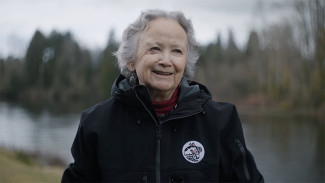 Older adult in a black jacket standing beside a calm lake, with trees and an overcast sky in the background.