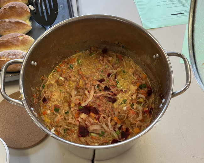 Large metal pot filled with a chunky soup sitting on a table beside a tray of bread rolls.