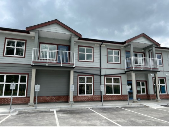Front section of a new two-storey grey and white apartment building with brick siding. A parking lot is in front of the entrance, featuring a short wheelchair ramp and ample accessible parking spaces.