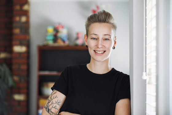 A young, smiling non-binary person with short hair, tattoos, and piercings leans against a wall.
