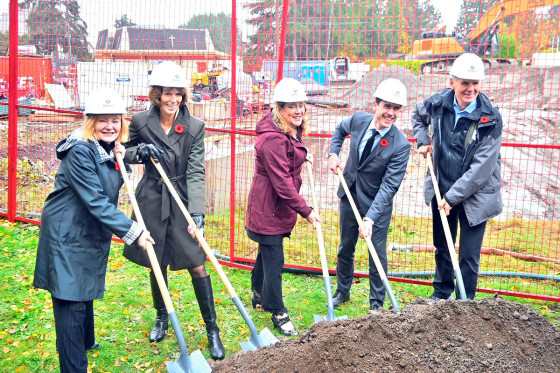 Five representatives wearing white hard hats place shovels into a pile of dirt at a groundbreaking event, with a construction site in the background.