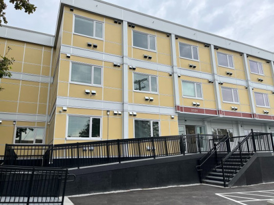 Front entrance of Aster Place, a new three-storey, flat-roofed yellow and grey apartment building. A black wheelchair ramp with railings and stairs frames the glass front door.