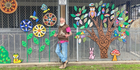 Smiling older man with a long white beard stands with thumbs up in front of a chain-link fence covered in garden art.