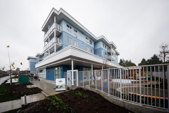 A modern, three-story building with light blue siding and white trim, featuring multiple balconies and large windows. The structure is surrounded by a metal fence, landscaped garden beds, and a paved walkway under an overcast sky.