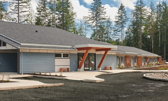 A single‑storey building with wood accents sits among tall trees under a bright sky.