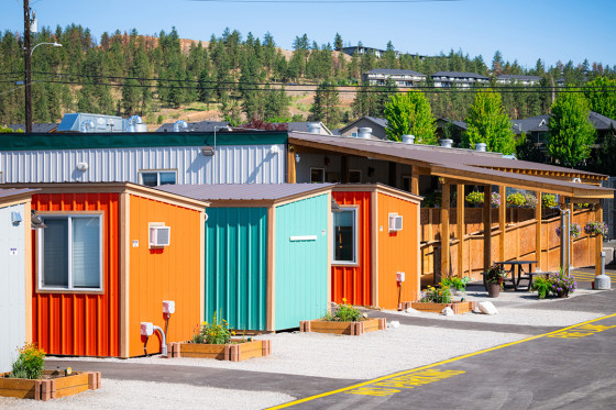 Row of colourful modular housing units with a covered walkway, set against trees and hillside in Kelowna.