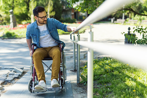 A wheelchair user reaches for a metal handrail along a paved park path bordered by grass and trees.