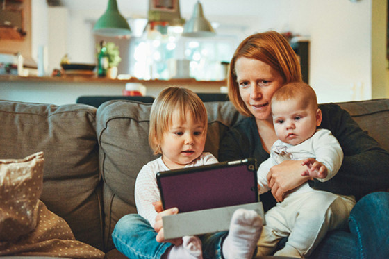 An adult sits on a sofa with two small children, holding a tablet together in a cozy living room.