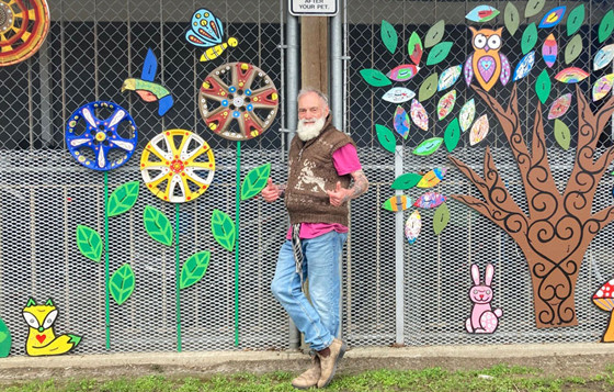 smiling older man with long white beard stands with thumbs up in from of chain-linked fence covered in garden art