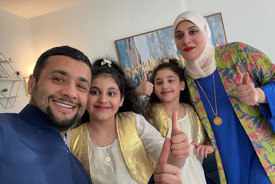 Inside a bright room, a family of four smiles at the camera with thumbs up. Mother and two daughters wear traditional Kurdish clothing in blue and yellow.