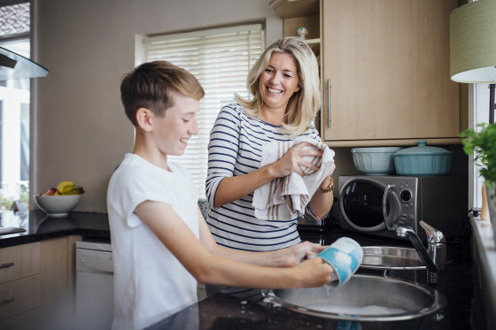 A mother and son are standing by a sink in the kitchen. The son is washing a cup and the mother is using a kitchen towel to dry the dishes. 