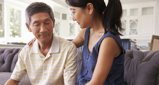 Daughter sits with her elderly father on a couch. Her arm is around his back and other hand is resting on her father's arm in his lap.