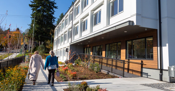 Two people walking along a sidewalk next to a modern residential building with landscaped gardens under clear blue skies in the fall.