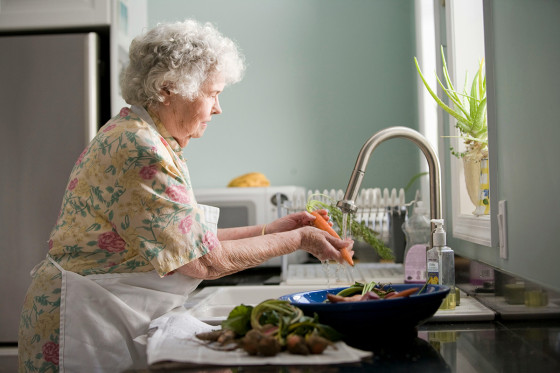 An elderly woman wearing a floral blouse and apron washes fresh carrots at the kitchen sink, preparing vegetables in a sunlit kitchen.