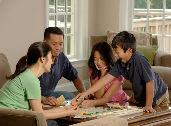 A smiling family of four sits in a bright living room playing a board game together, enjoying quality time around a coffee table.