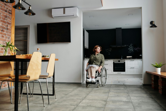 A person in a wheelchair is centered in a modern kitchen and dining area with white cabinets, a black backsplash, stainless steel appliances, a wooden dining table with four chairs, and a wall-mounted air conditioner above a flat-screen TV; a potted plant rests on a wooden bench to the right.