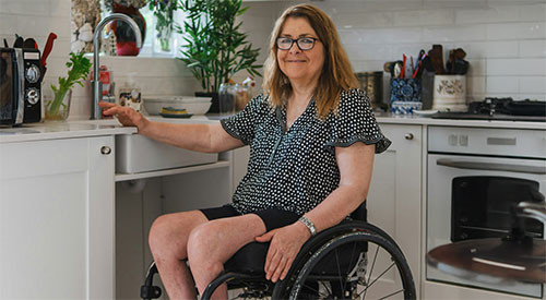 A person seated in a wheelchair in a modern kitchen, positioned near white cabinets and a countertop with various items, including a coffee maker and potted plant. The background features an oven and additional kitchen essentials.