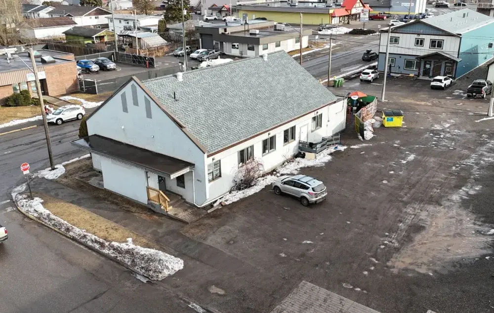 Angled aerial view of a small building with a parking lot, nearby homes, patches of snow, and surrounding streets in a town.
