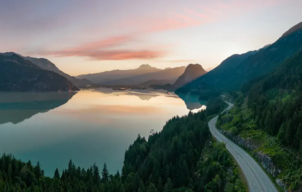 A calm river reflecting mountains at sunset, with a winding road along a forested hillside under a pink-and-blue sky.